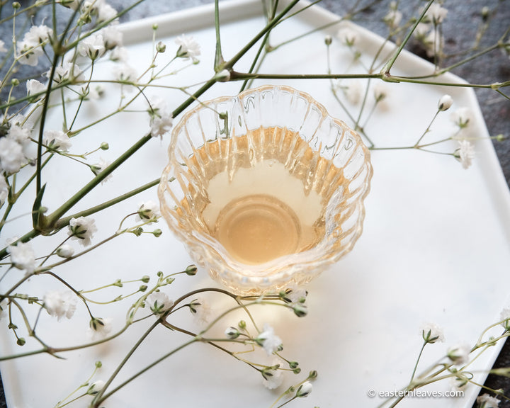 Chinese tea cup in glass with bright infusion of green tea, white tiny flowers on white ceramic tray from Dehua, China